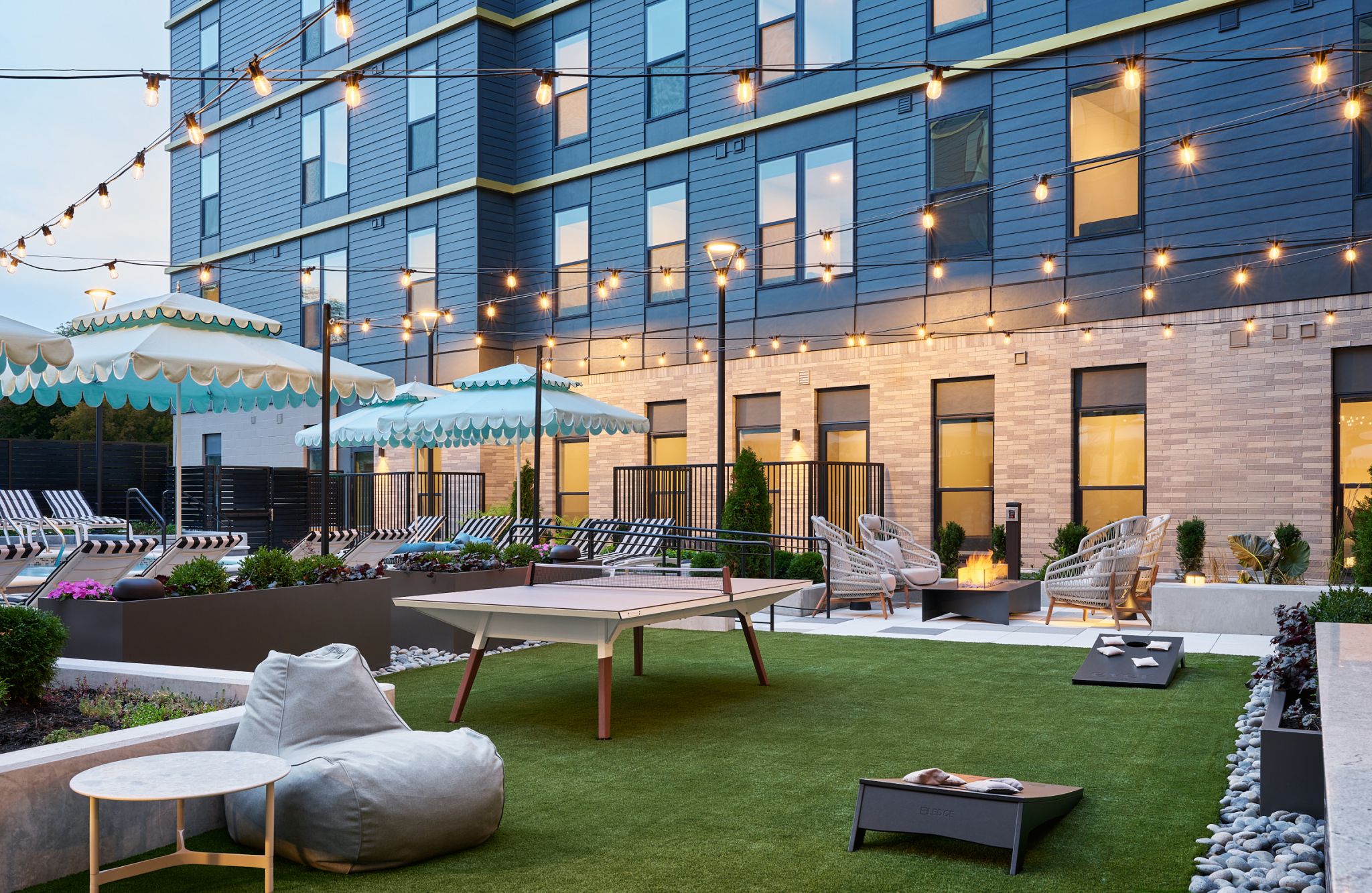 Outdoor courtyard with string lights, lounge chairs, umbrellas, ping pong table, and cornhole game on green turf.