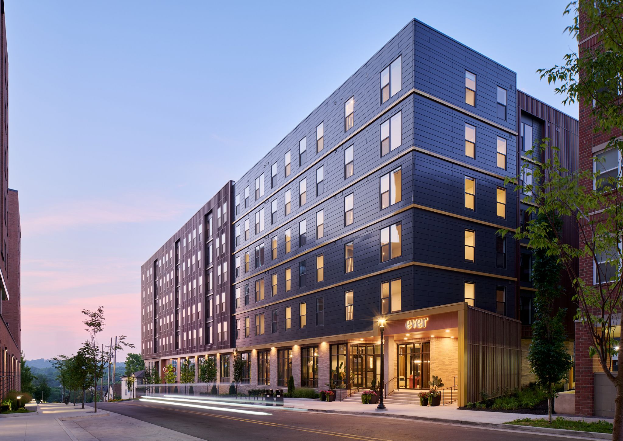 Modern mid-rise apartment building with large windows, lit warmly at dusk, on a quiet city street.