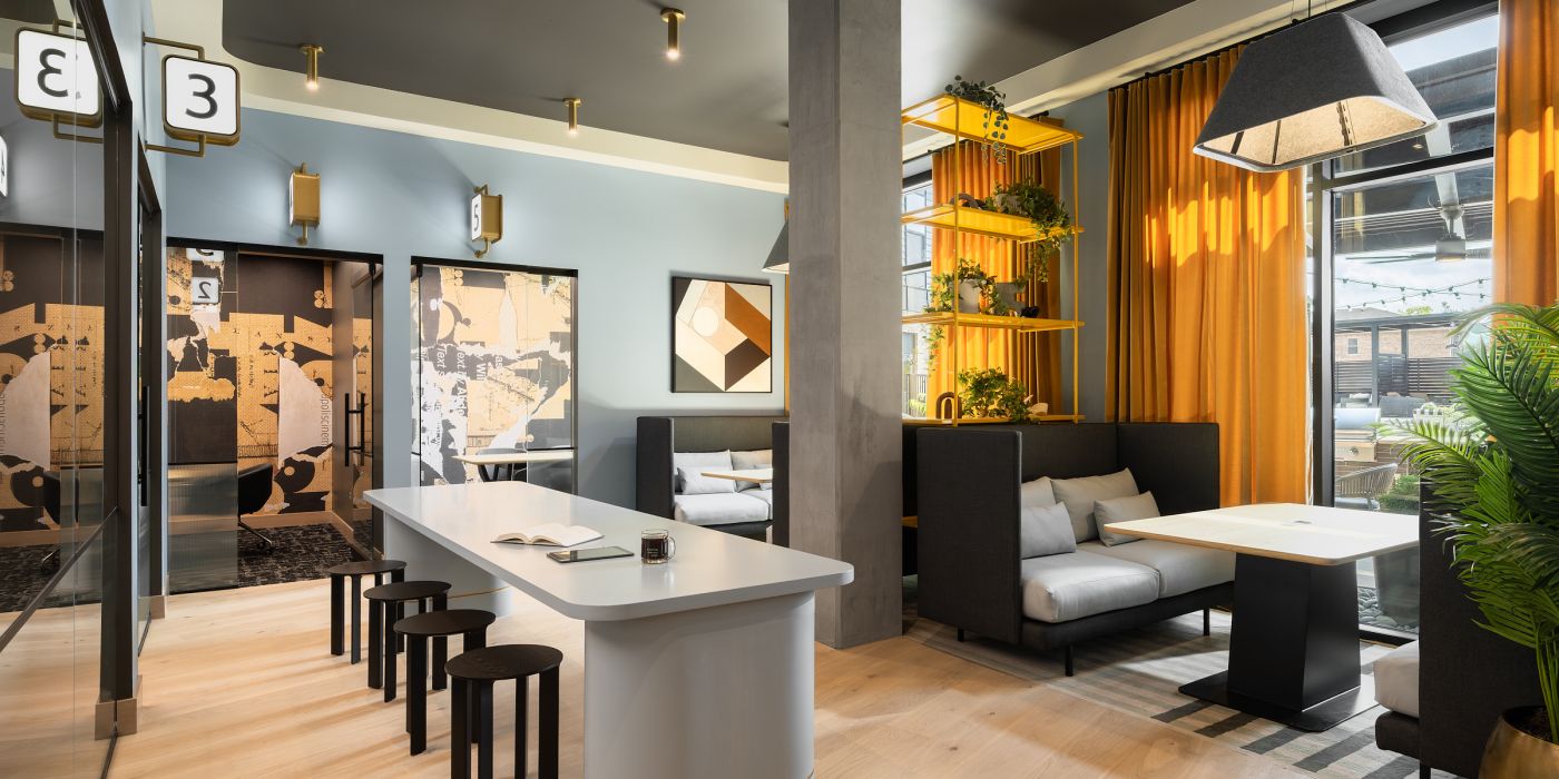 Espacio de coworking moderno con una mesa larga, taburetes, reservados, plantas y grandes ventanales con cortinas amarillas.