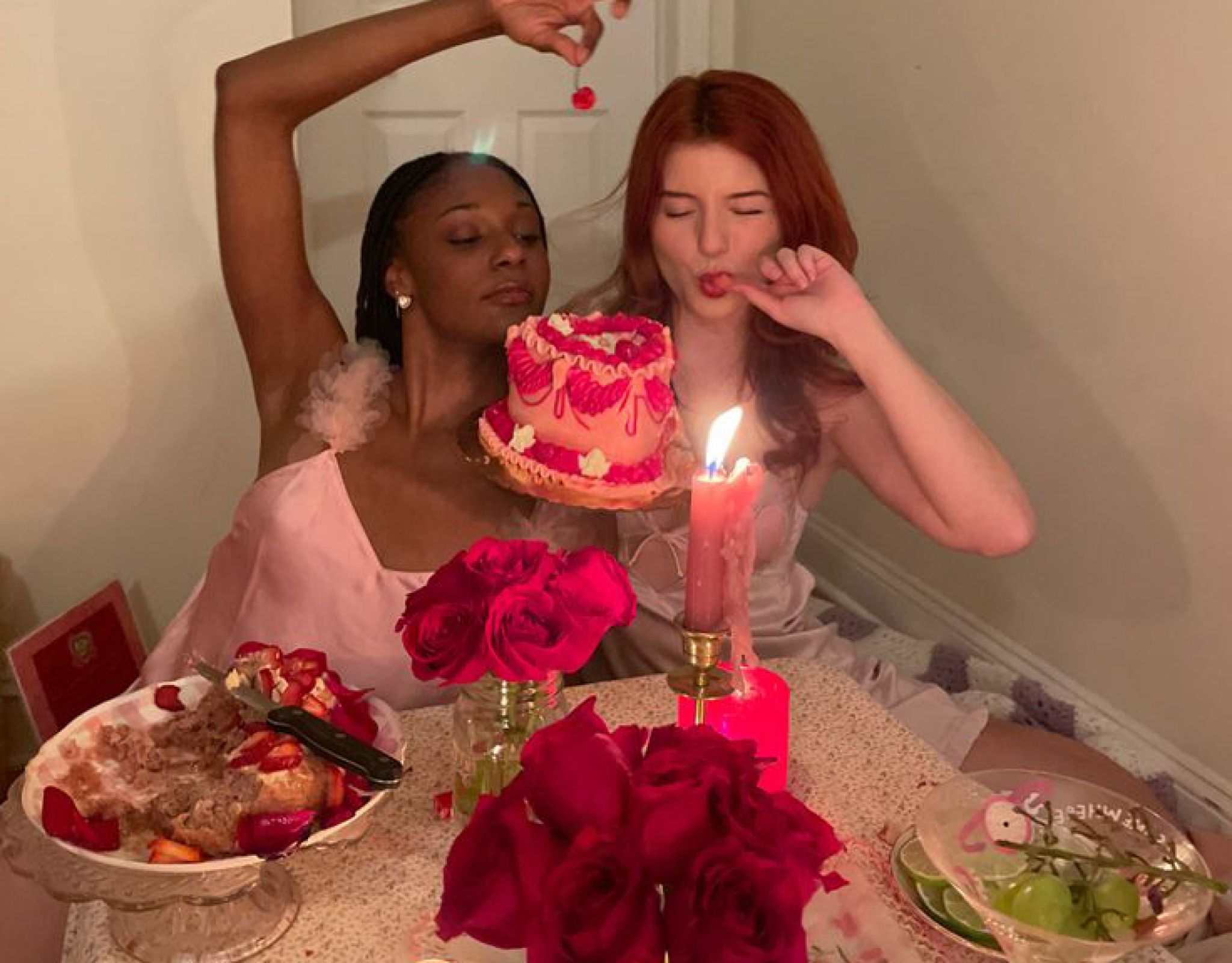 Two women in pink dresses at a decorated table with a cake, roses, and candles, giving playful expressions this February in West Lafayette.