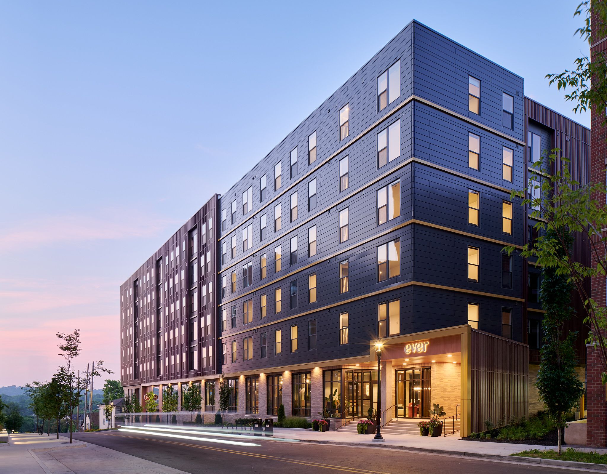 Modern mid-rise apartment building with large windows, lit warmly at dusk, on a quiet city street.