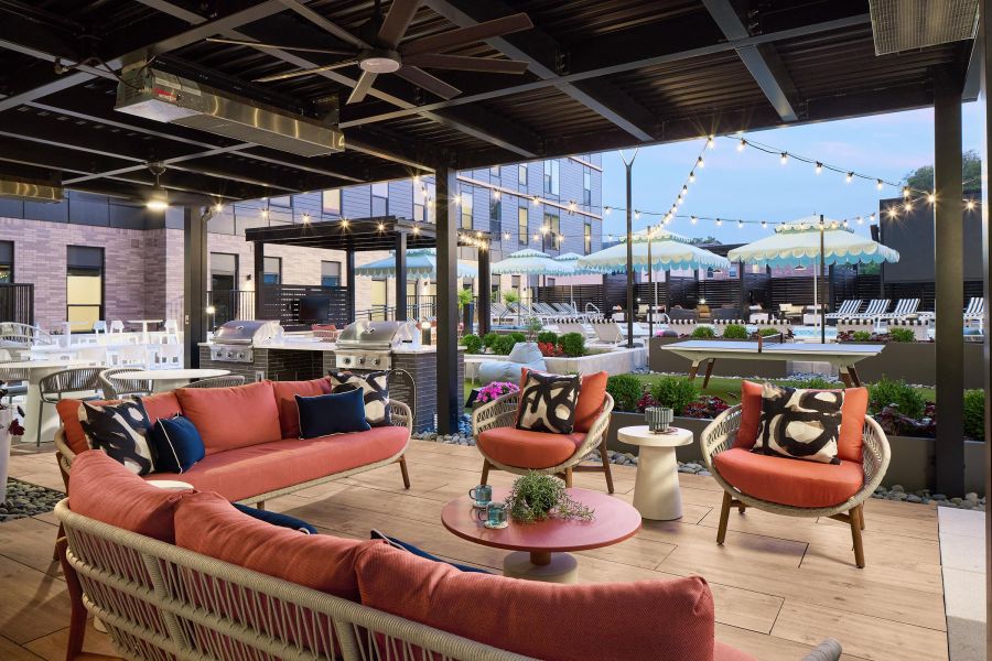 Ever West Lafayette outdoor lounge with coral sofas, chairs, string lights, and a view of a pool with umbrellas in the background.
