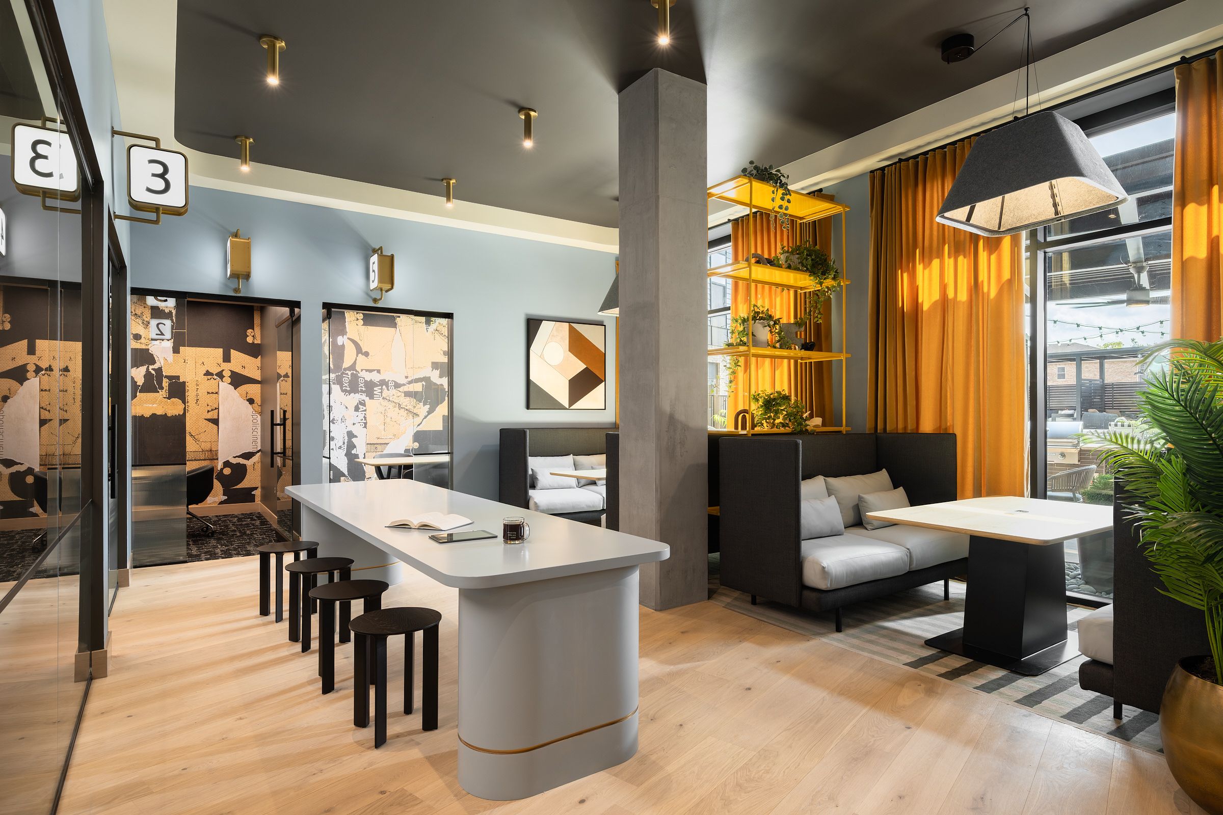 Espacio de coworking moderno con una mesa larga, taburetes, reservados, plantas y grandes ventanales con cortinas amarillas.