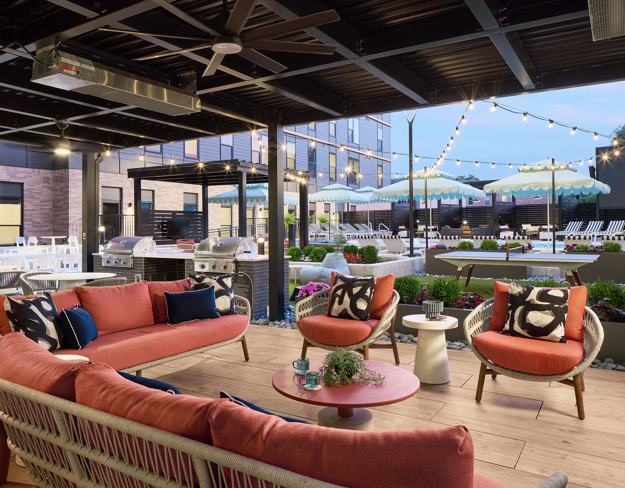 Outdoor patio with pink seating, string lights, and pool area with striped umbrellas in the background.