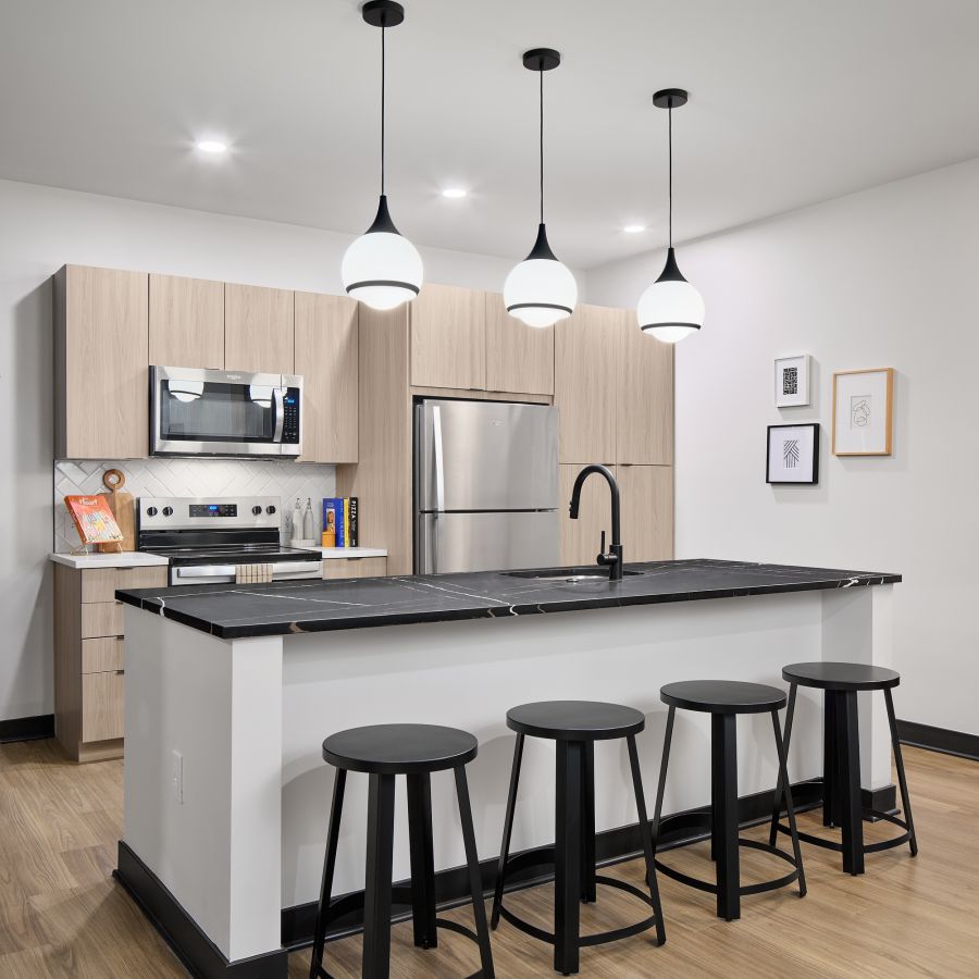 Modern kitchen with light wood cabinets, black island, four black stools, and three pendant lights overhead.