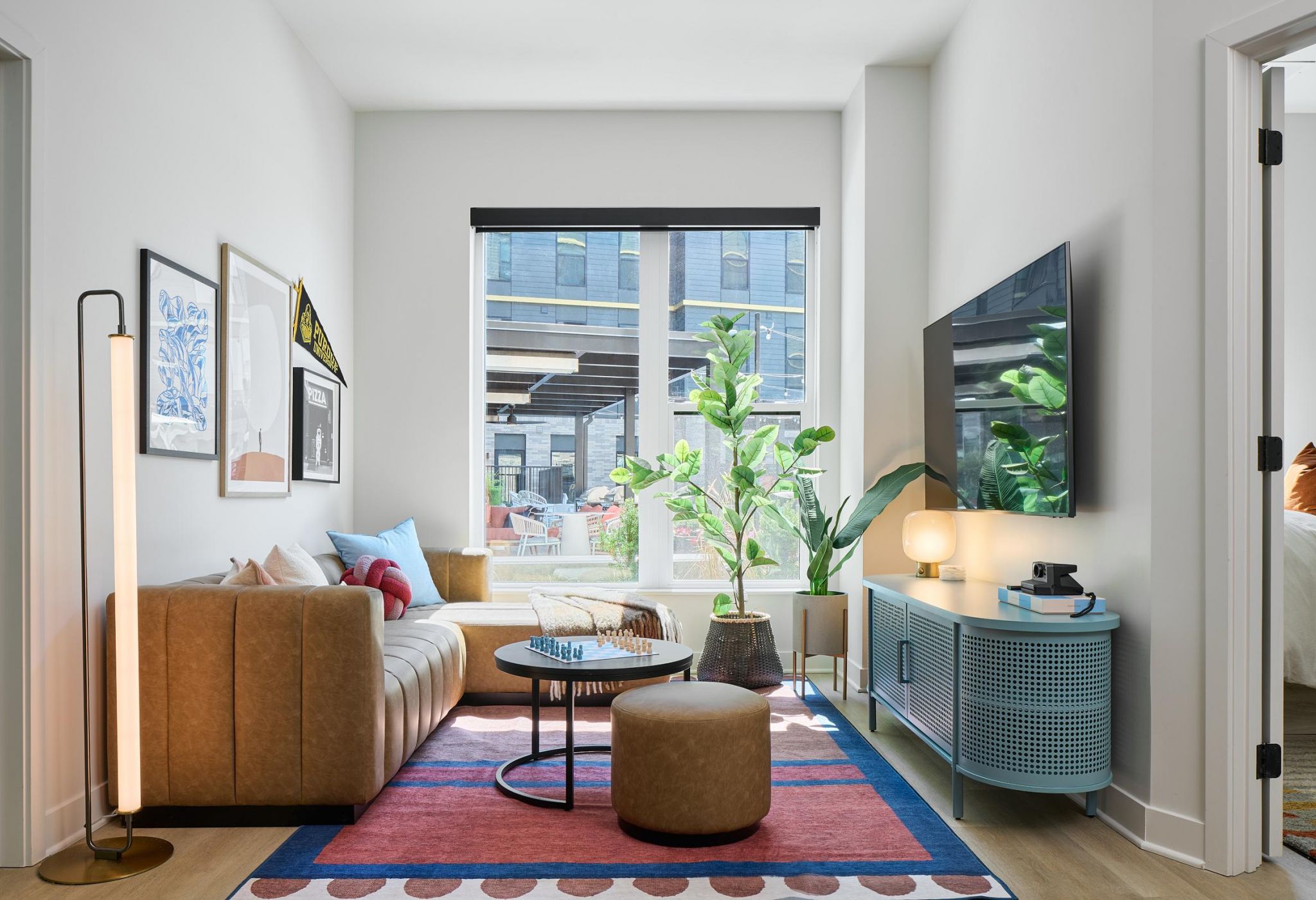 Ever West Lafayette modern living room with tan sofa, colorful rug, plants, wall art, and TV on the wall near large windows.