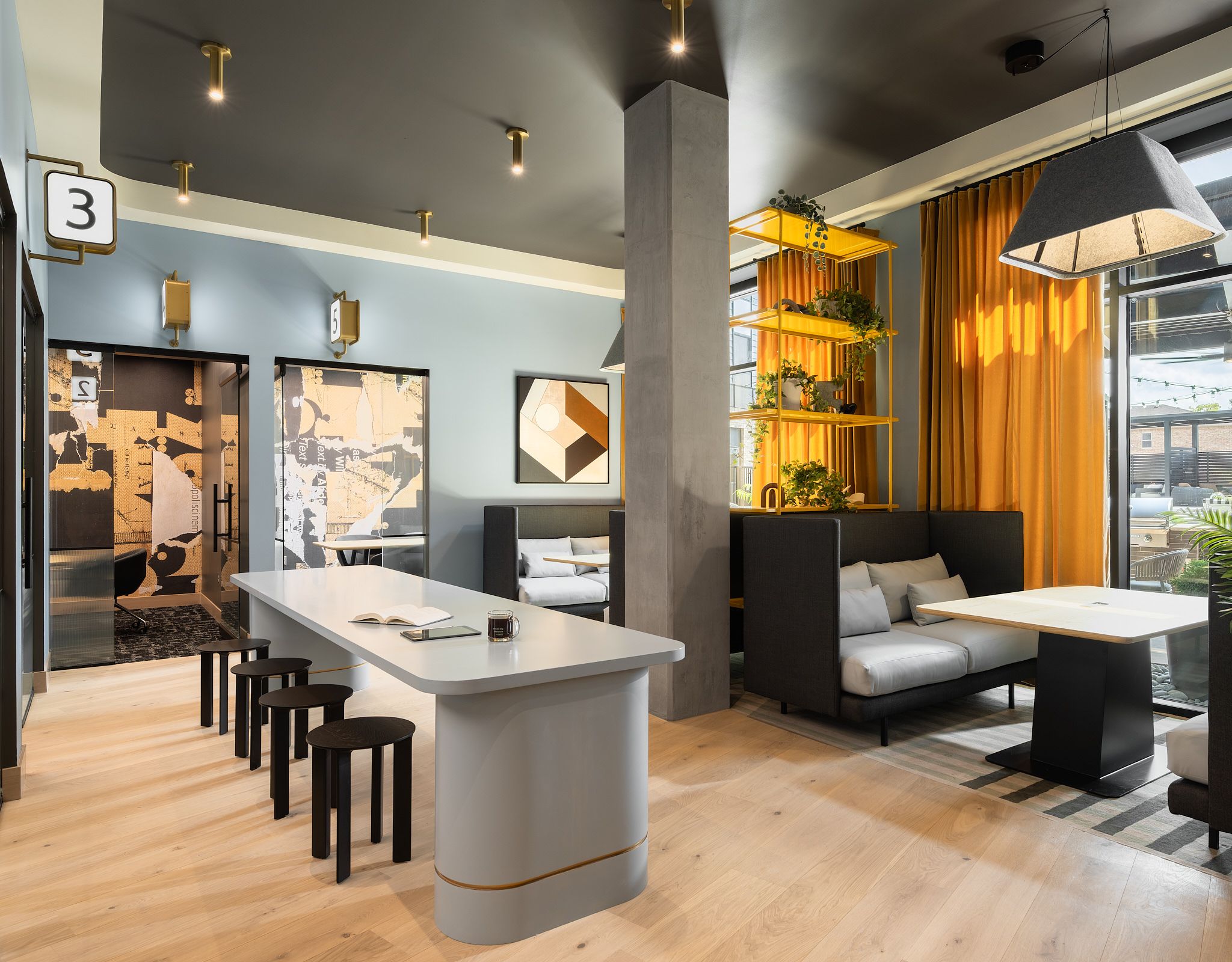Modern coworking space with long table, stools, booth seating, plants, and large windows with yellow curtains.