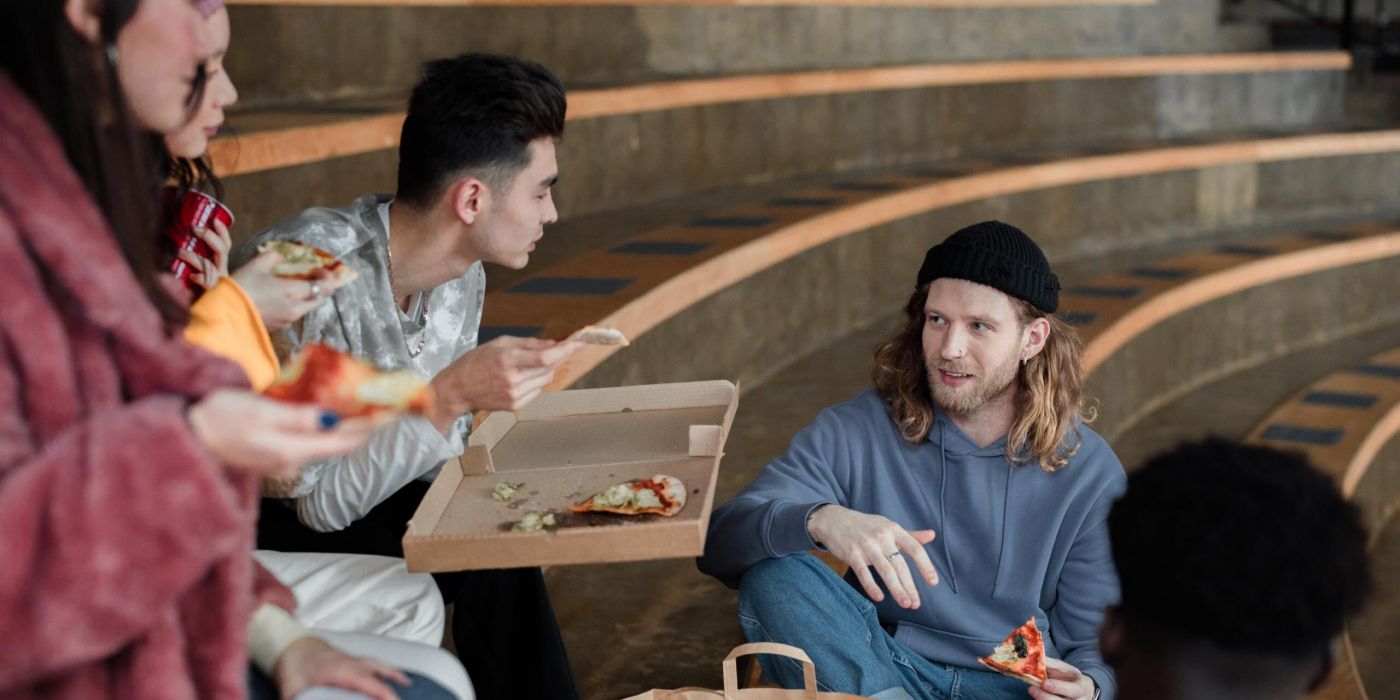 Two people sit on steps, eating pizza from a box and talking. One has a beanie; the other holds a slice of pizza.