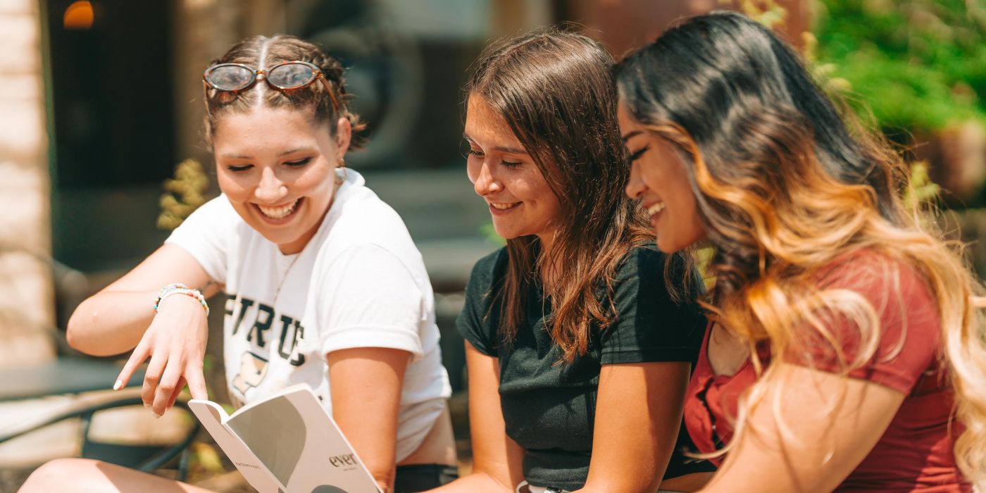 Three female college students sitting outside and looking at a catalog together