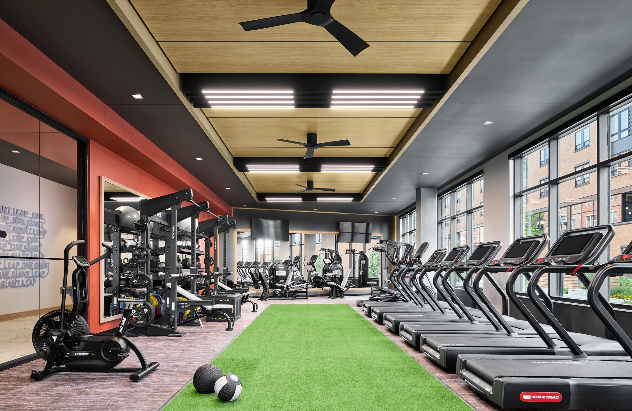 Modern gym with treadmills, weight machines, a green turf strip, exercise balls, and large windows.