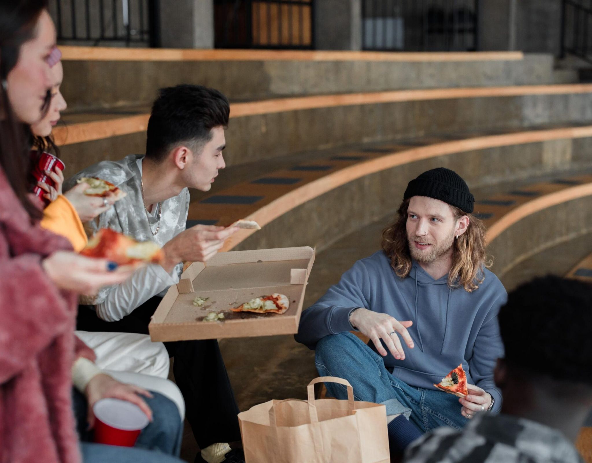 Two people sit on steps, eating pizza from a box and talking. One has a beanie; the other holds a slice of pizza.
